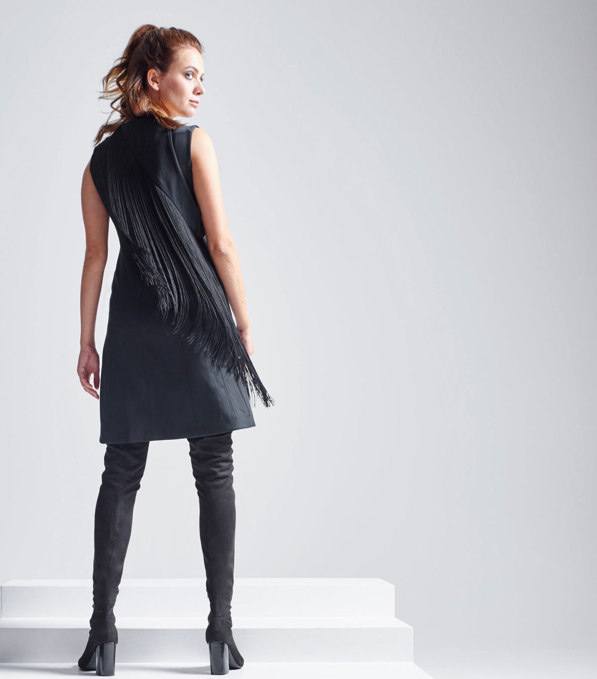 Das Model trägt den abnehmbaren Multiplyfashion Fransenkragen und das Multiplyfashion Etuikleid in Kombination mit Overknee Stiefeln. Rückenansicht während die Fransen in der Bewegung schwingen.